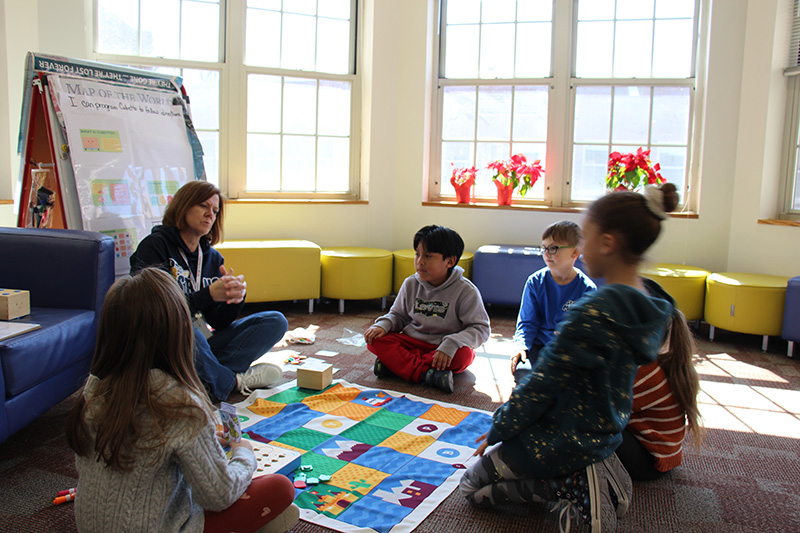 First grade students squat on the floor, with a large mat in front of them. There is a woman with shorter hair working with them.They are programming a small robot.