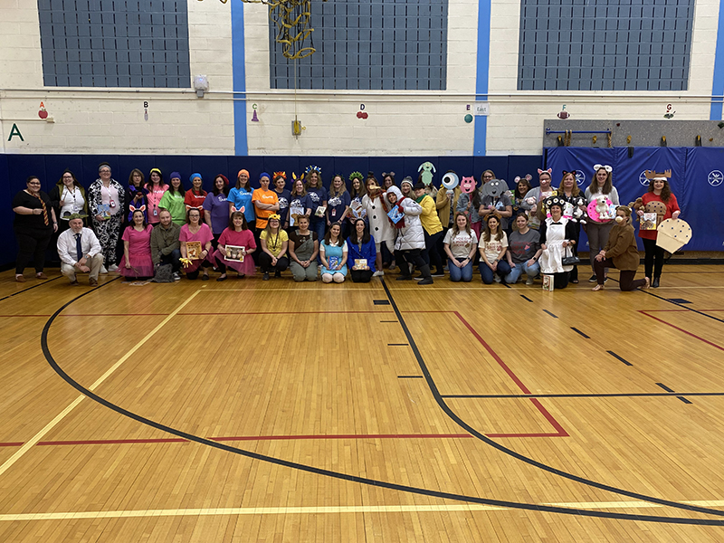 A large group of adults stands together all dressed as literary characters in a gymnasium.