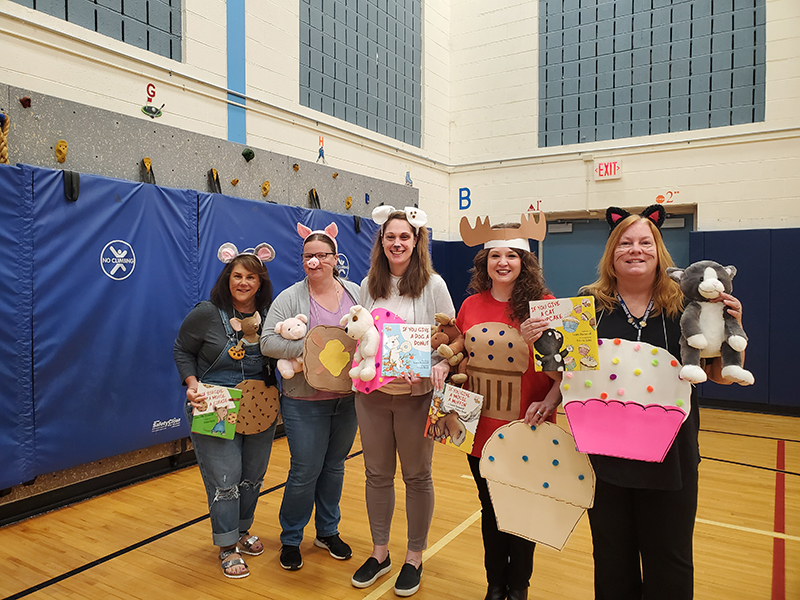 Five women stand with large cutouts of cupcakes and cookies. They have animal ears on them.