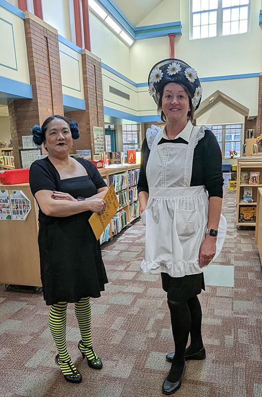 Two women dressed in costume stand together and smile. The one on the left is dressed in a black dress with yellow and black tights. The woman on the right is dressed with a flowered hat, black dress and white apron.