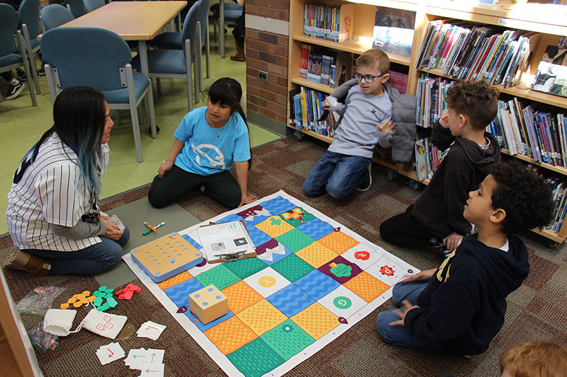 Four first grade students sit around a mat with patterns on it. There is a woman with long dark hair kneeling next to them.