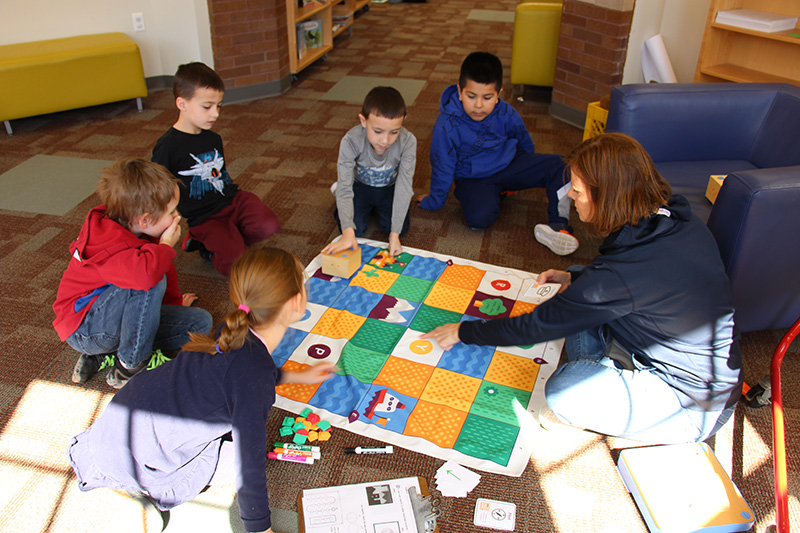 First grade students squat on the floor, with a large mat in front of them. There is a woman with shorter hair working with them.They are programming a small robot.