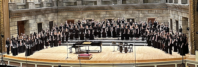125 high school students stand on risers and perform at a large hall.