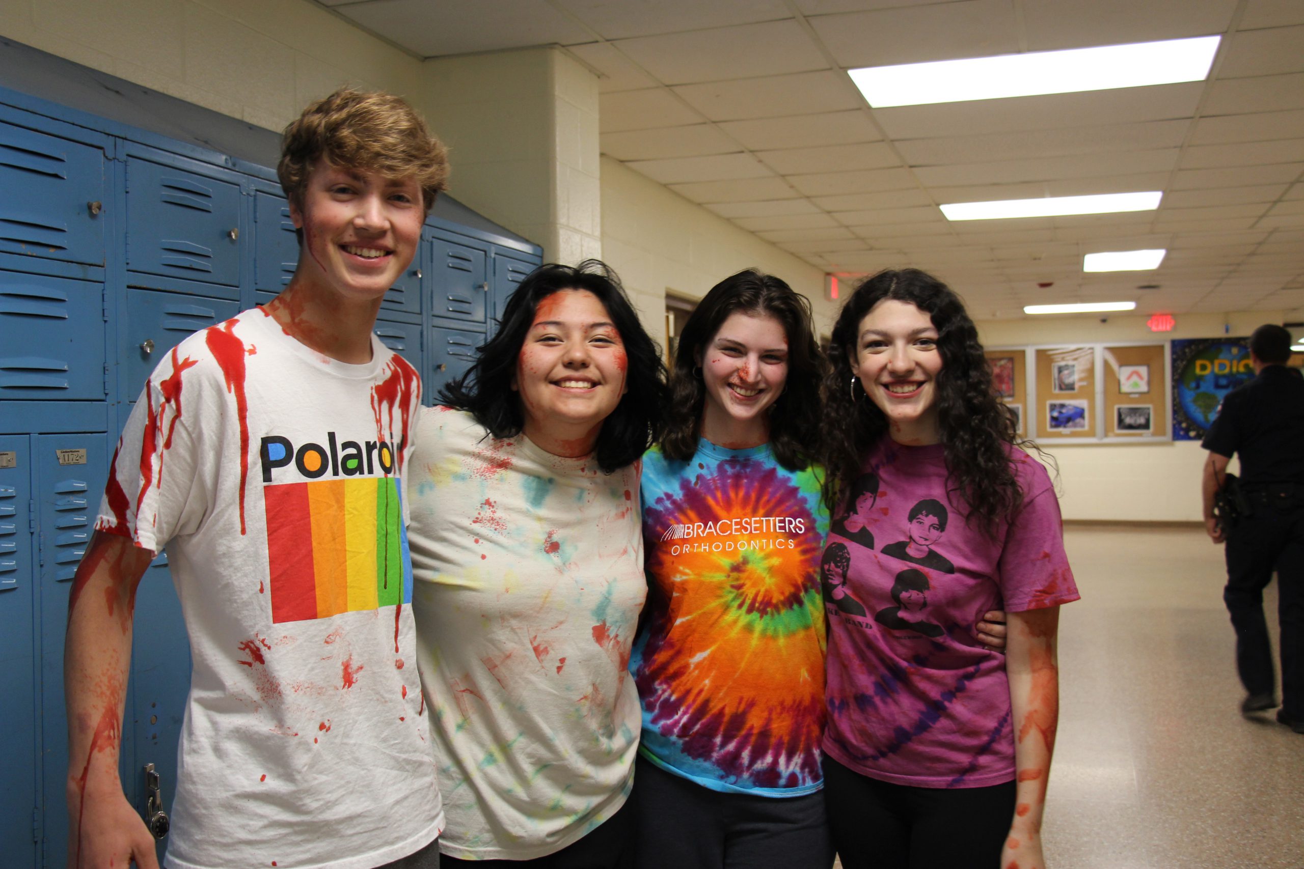Four high school students, one young man and three young women, stand smiling. They have fake blood on them.