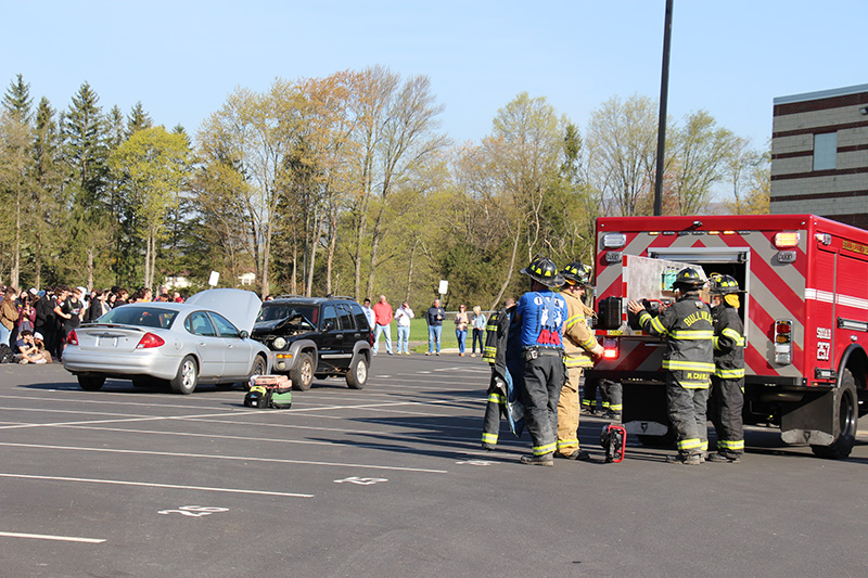 Emergency vehicles on the right with two cars in the middle that have collided. A group of high school juniors are along the side watching this mock drill.