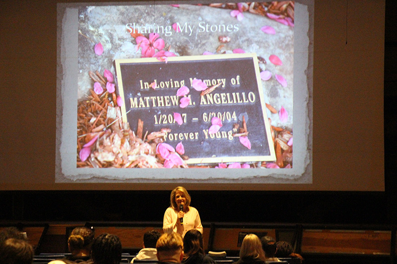 A woman is in front of an auditorium filled with high school kids. Behind her is a screen with a picture of a gravestone . Engraved is In loving memory of  Matthew M. Angelillo 1/20/87 -6/20/04 Forever Young