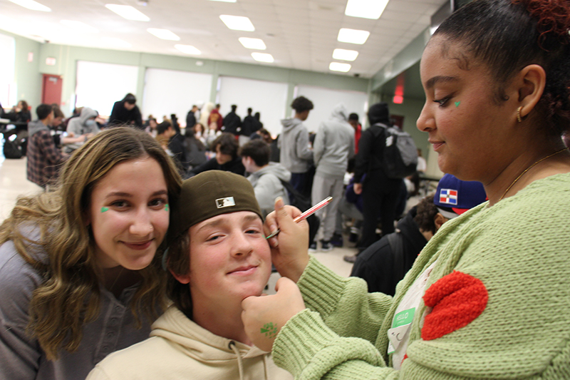 A high school girl dressed in a green sweater paints on a high school kid's face. Another high school girl smiles next to him.