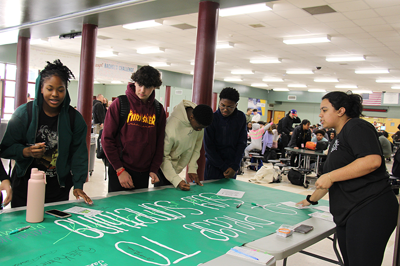 A group of four high school students look at and sign a large green poster that says I promise to Say Something.