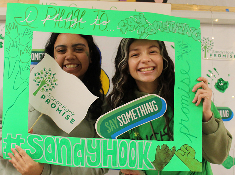 Two high school students, both girls, smile as they hold up a green frame. They have signs that says "Say Something" and Sandy Hook Promise.