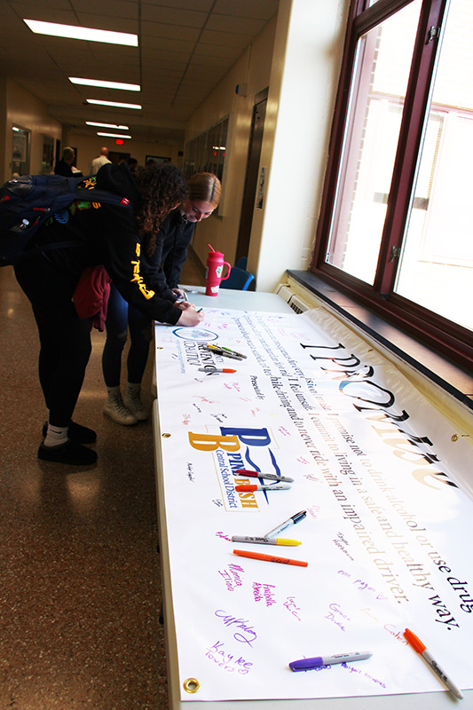 A long table with a white banner on it. The banner says I Promise with a Pine Bush blue and gold logo , a Tri-County prevention logo and many signatures.