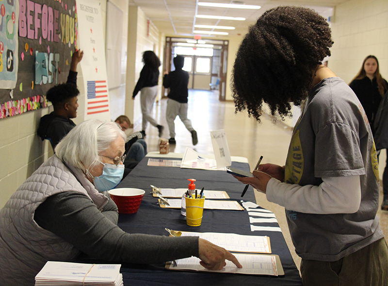 A high school student with curly dark hair leans over a table and  fills out a form on a clipboard. A woman with white hair points to something ona piece of paper.