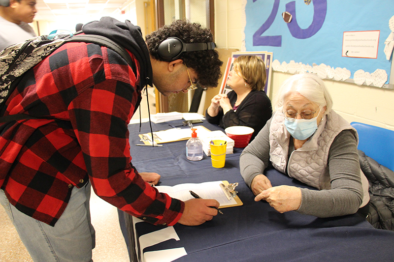 A high school student with dark curly hair wearing a red and black check shirt fills out a form while a woman with white hair helps him out.