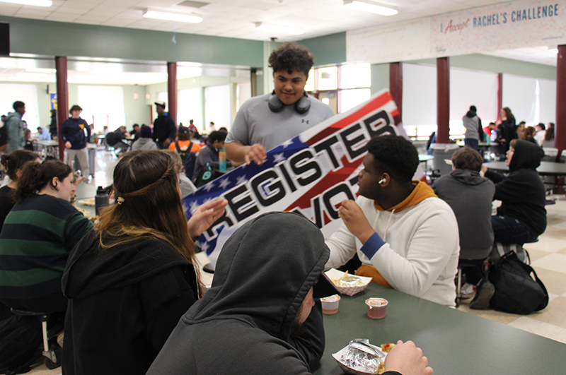 A high school student carries a red, white and blue sign that says Register to Vote as he talks to three students at a table.