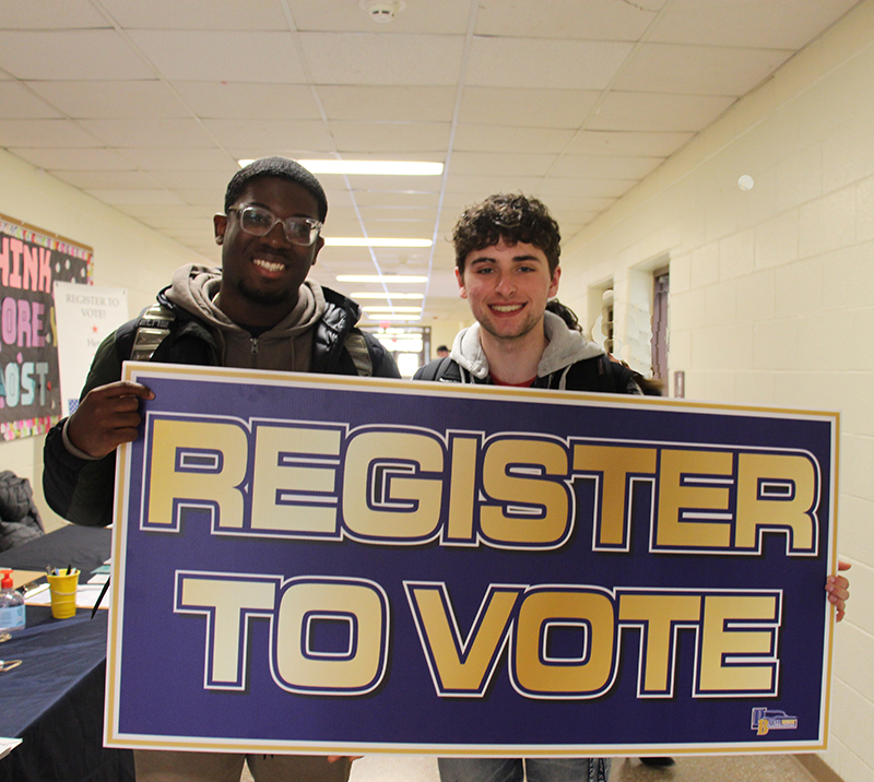 Two high school students smile as they hold a blue and gold sign that says Register to Vote.