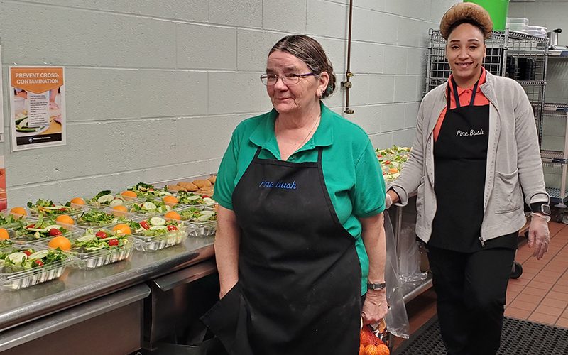 Two women, both with black aprons, stand by many prepared salads that are sitting on the counter next to them.