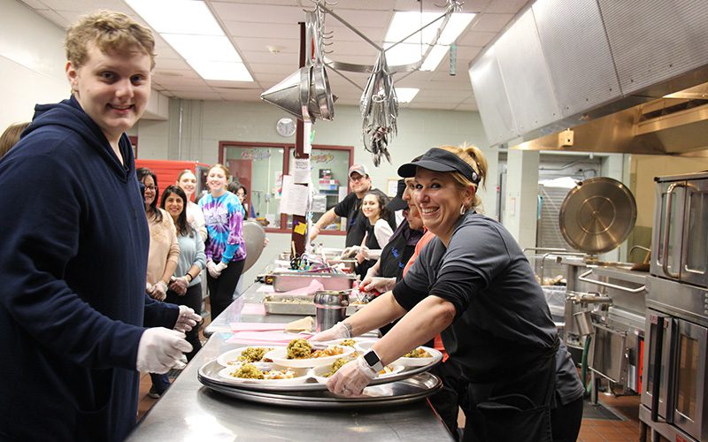 There are people on the right and left side of this long counter. The people on the right are loading food up on the trays on the counter while the people on the left wait to take the trays.