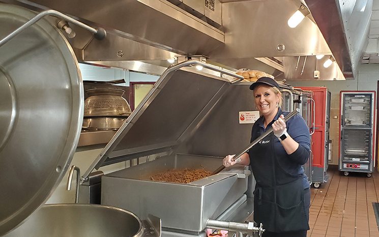 A woman in a black apron, blue shirt and her hair pulled back, uses a very large utensil to stir a huge pot of meat cooking.