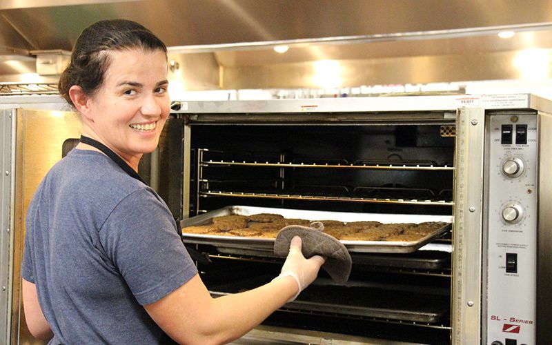 A woman with her hair pulled back, wearing a blue short sleeve shirt, takes a tray of hamburgers out of the oven. She is looking over her shoulder and smiles at the camera.