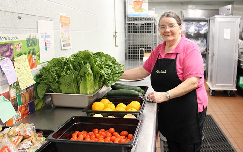 A woman in a pink shirt and black apron smiles with lots of vegetables in front of her, lettuce, tomatoes, cucumbers.