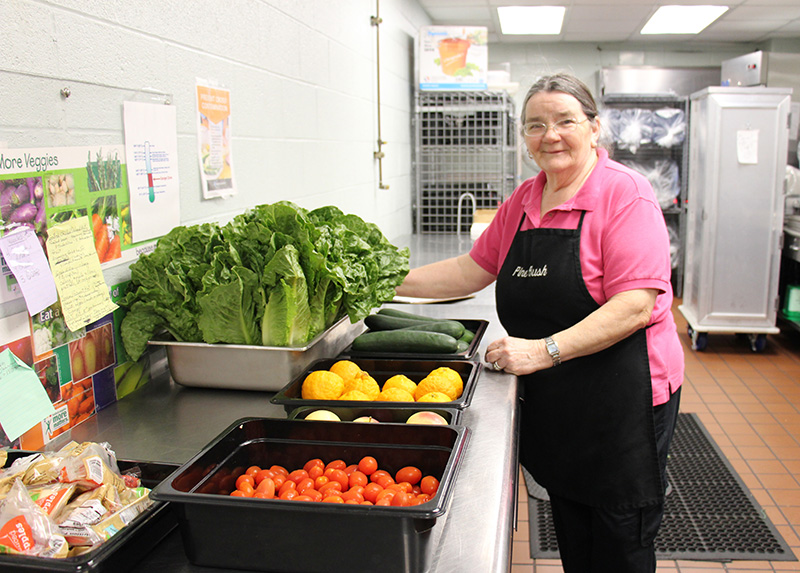 A woman in a pink shirt and black apron smiles with lots of vegetables in front of her, lettuce, tomatoes, cucumbers.