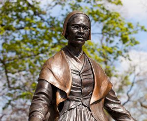 A blue sky in back, green tree and a bronze statue of a woman wearing a shawl, wire rim glasses and a hat.