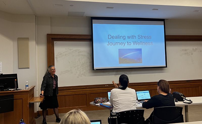 A large screen at the front of a classroom says Dealing With Stress Journey to Wellness. There is a woman at the front of the classroom talking to those seated.