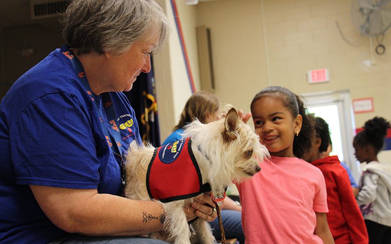 A girl in a pink shirt with her dark hair pulled back in a ponytail pets a little white dog who is being held by a woman wearing a blue shirt.