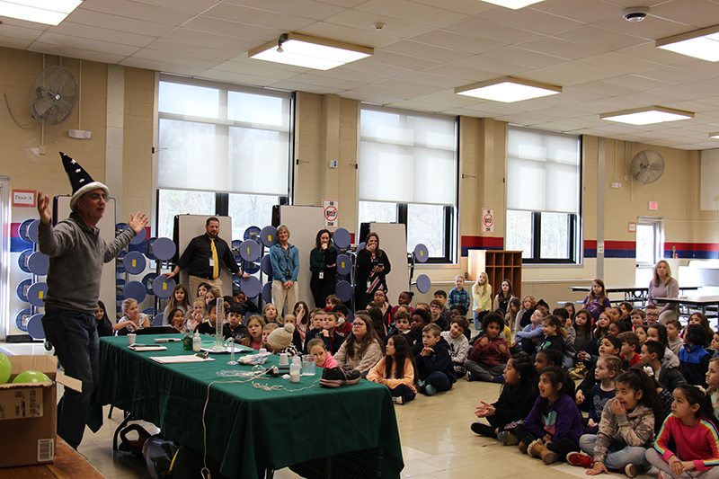 A large group of kids on the floor of a room with a man in front wearing a tall blue hat with stars on it.