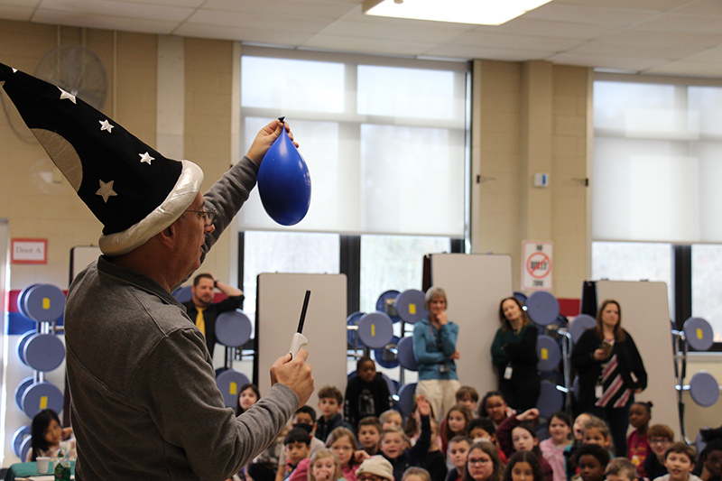 Students sit and watch as a man with a tall blue hat with stars on it holds a flame to a blue balloon.