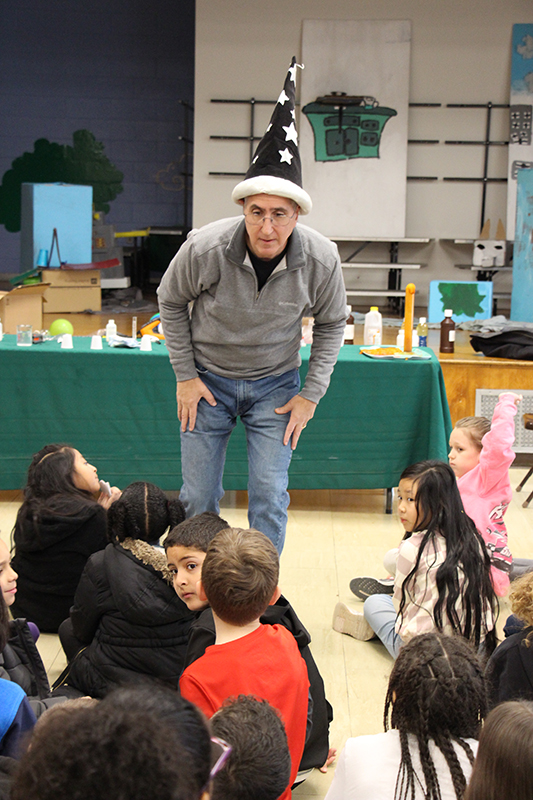 A man with a gray shirt and wearing a tall blue hat with white stars on it leans  in to a group of kids sitting on the floor. He is answering questions for them.