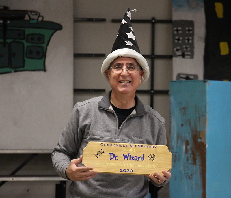 A man wearing a gray shirt, holds a wooden plaque up and he is smiling. He is wearing a tall blue hat with white stars on it.