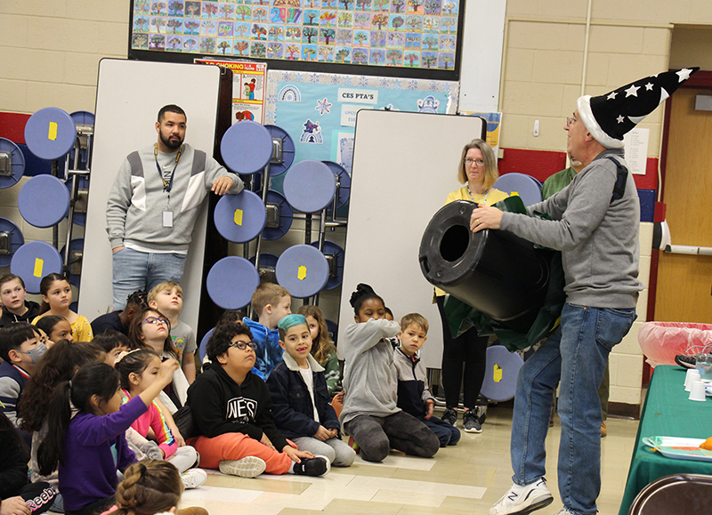 A man stands in front of a large group of kids sitting on the floor. He is holding a black trash can that has a hole in the bottom and the top covered in material.