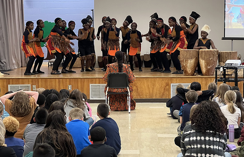 A view from the back of the room with rows of elementary students watching the stage where a large group of kids dressed in traditional African garb are playing drums.