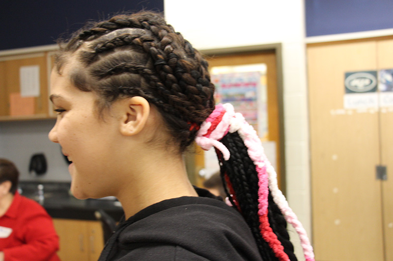 A fifth-grade girl with long grades. She has them pulled back into a large ponytail with pink and white crocheted hair ties. We are viewing from the side.