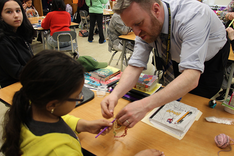 A man with short hair leans over a desk to help a fifth-grader with crocheting.