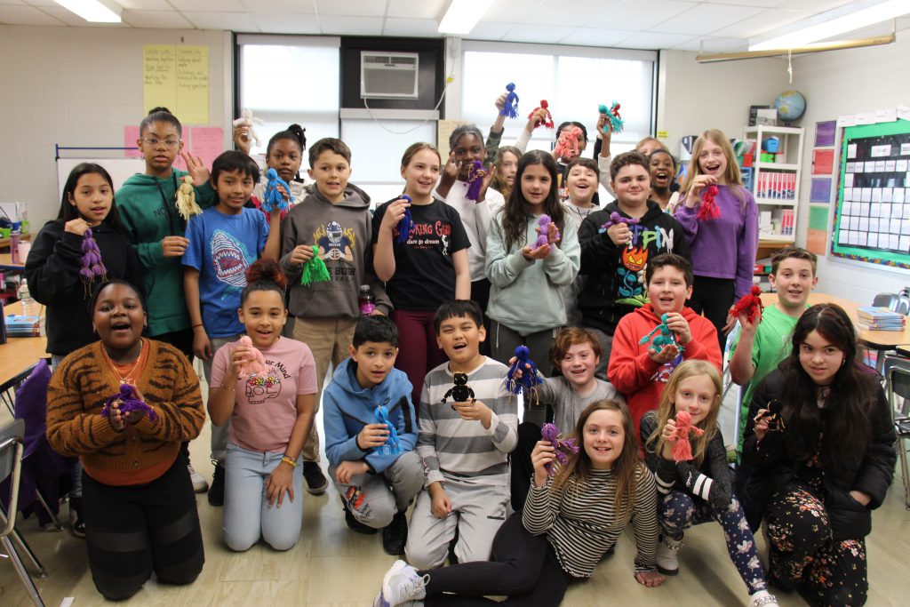 A large group of fifth-graders stand together holding up their dolls that are made of yarn.