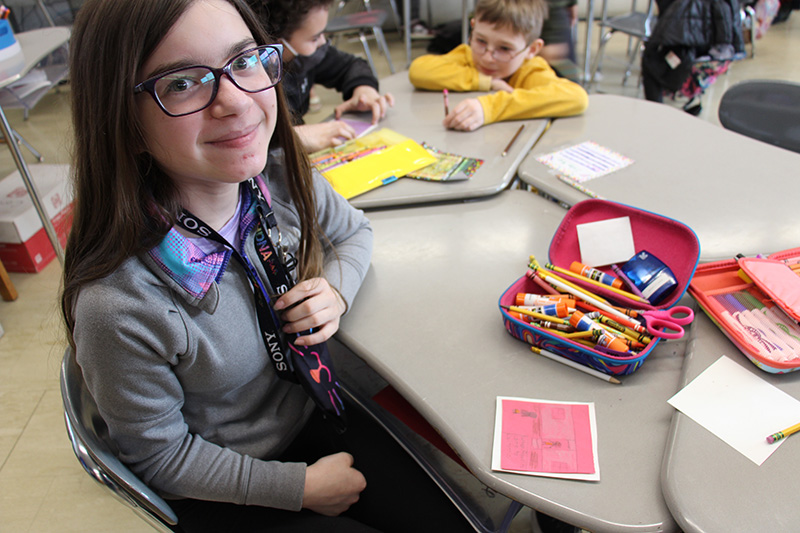 A fifth-grade students sits at a desk with colored pencils and crayons. She colored a square that is red. She is wearing glasses and is smiling.