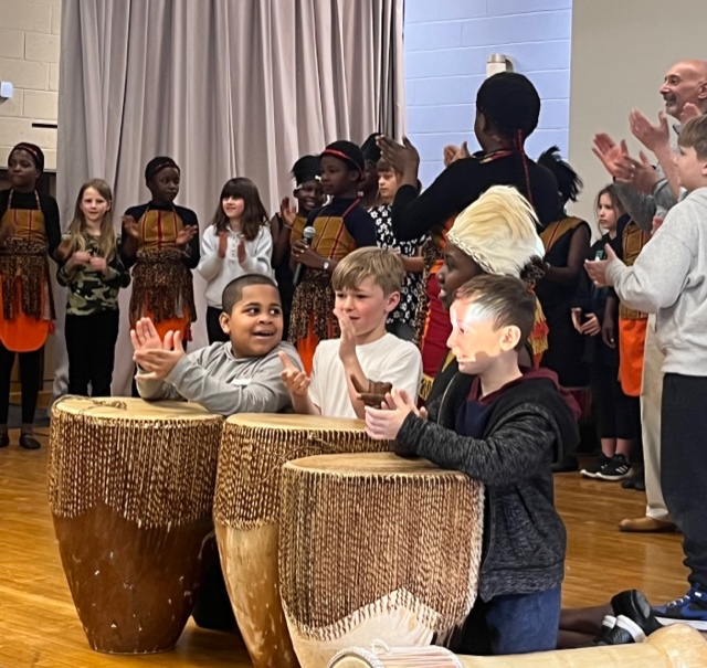 In the background of kids dressed in traditional African garb . In front are elementary school students playing drums and smiling.