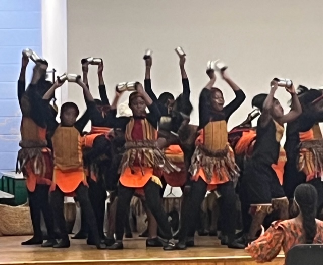 A large group of kids dressed in traditional African garb hold small white instruments over their heads.