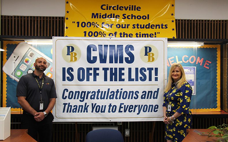 A man with a beard in a blue polo shirt stands on the left of a banner and a woman with long blonde hair wearing a blue and yellow printed shirt and pants stands on the right. There is a banner between them that says CVMS is off the list! Congratulations and thank you to everyone. Above the banner is another that says Circleville Middle School "100% for our students 100% of the time!"