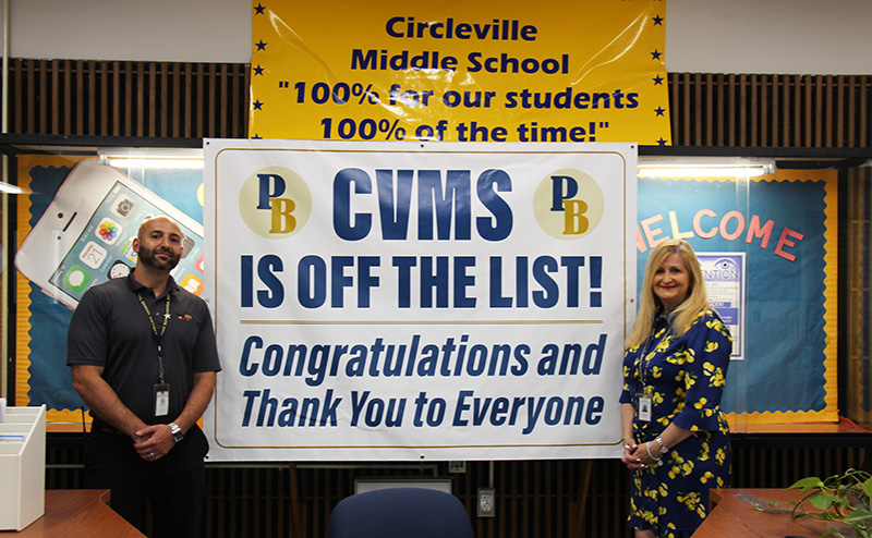 A man with a beard in a blue polo shirt stands on the left of a banner and a woman with long blonde hair wearing a blue and yellow printed shirt and pants stands on the right. There is a banner between them that says CVMS is off the list! Congratulations and thank you to everyone. Above the banner is another that says Circleville Middle School "100% for our students 100% of the time!"
