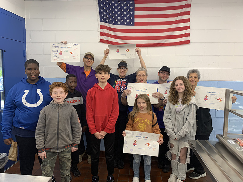 Six middle school kids and five adults who are Food services workers, stand together smiling. The adults ar eholding signs the kids made to honor them.