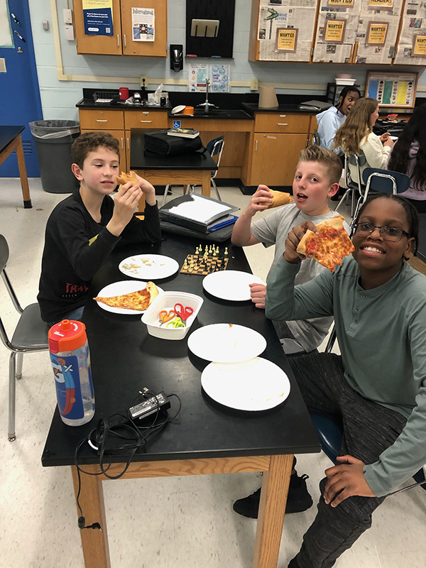 Three middle school boys sit around a table eating pizza. They are smiling.