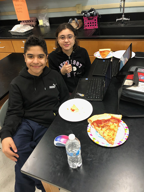 Two middle schools kids smile and there is pizza on the table.