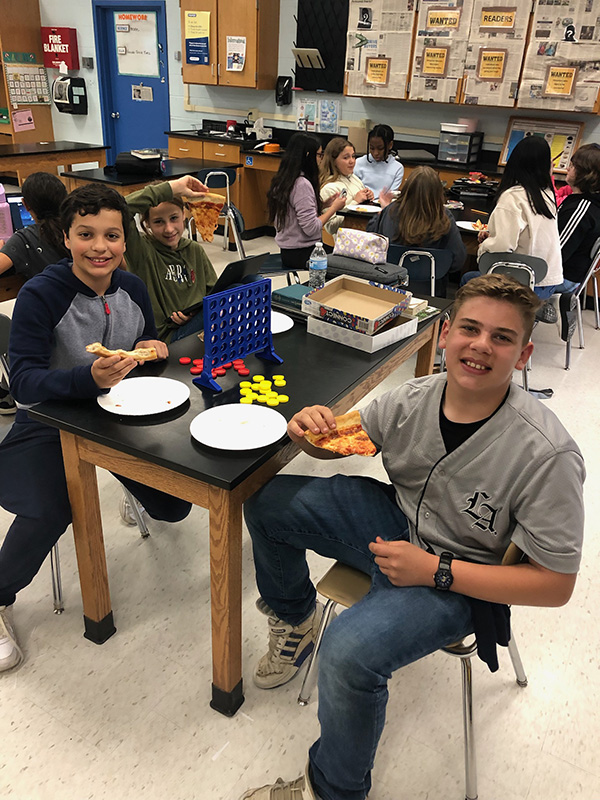 Two middle school boys smile at the camera. They have pizza in their hands and there are others in the background eating pizza.