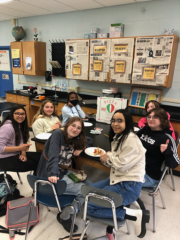 A group of 7 middle school students sit around a table eating pizza. they are smiling.