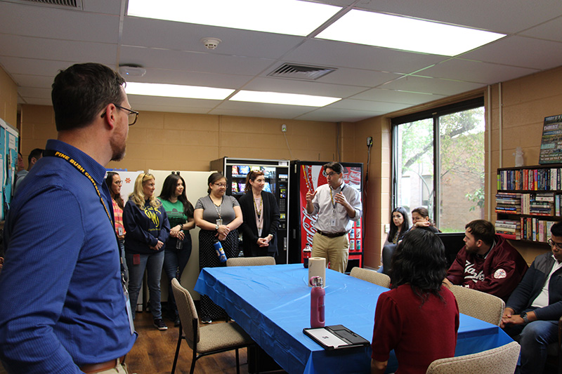 A group of people stand around the perimeter of a room. There is a table in the center with a blue table cloth. a man wearing a white shirt and khaki pants stands in front talking.