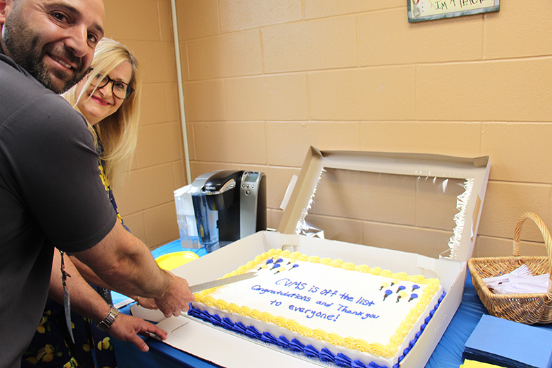 A large cake with blue and gold icing is on a table. It says CVMS is off the list Congratulations and Thank you to everyone! A woman with blonde hair and a man wearing a gray shirt are cutting it and smiling at the camera.