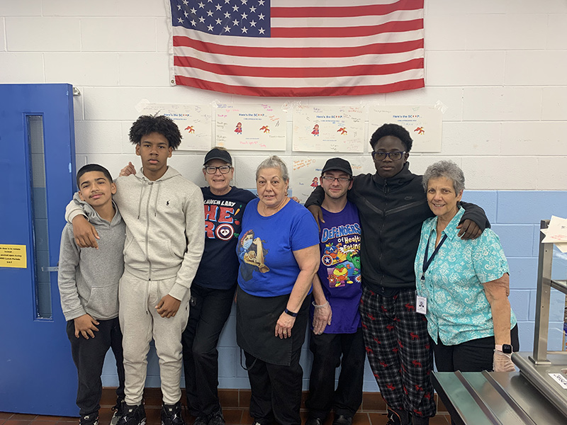 Four adults stand with three middle school students arm in arm. They are in a cafeteria. The background has an American flag and posters honoring the adults.