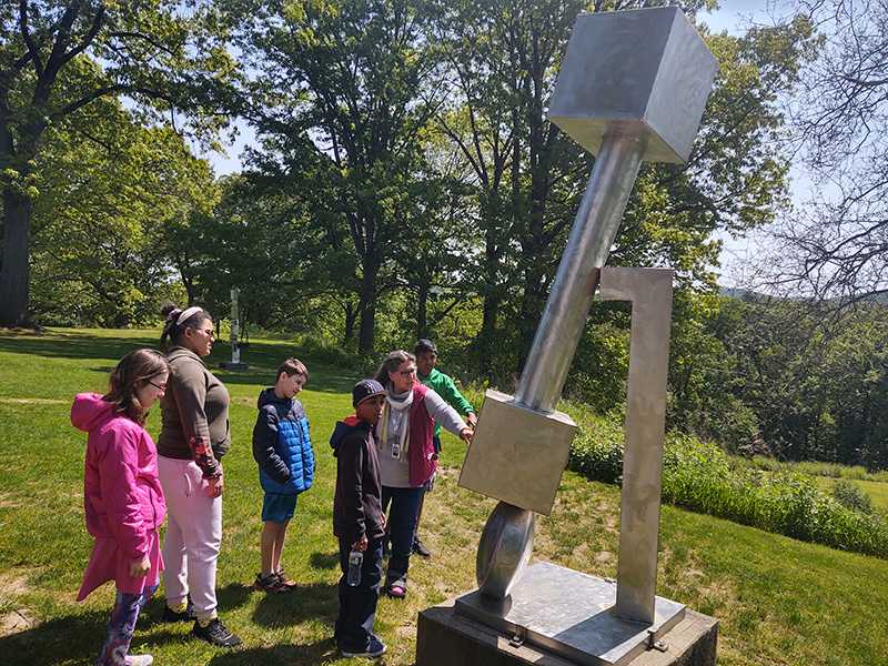 Four middle school children and an adult look at a large silver-colored sculpture. There are trees in the background on a sunny day.